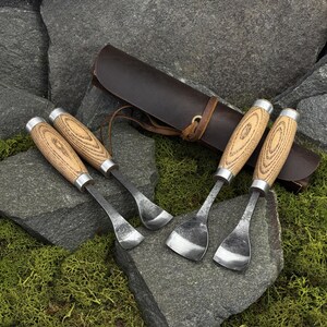 May include: A set of four woodworking tools with light-colored wood handles and silver metal ferrules. Two tools feature curved blades, while two have wider, flat-edged blades. A dark brown leather roll-up case with a leather tie is included.
