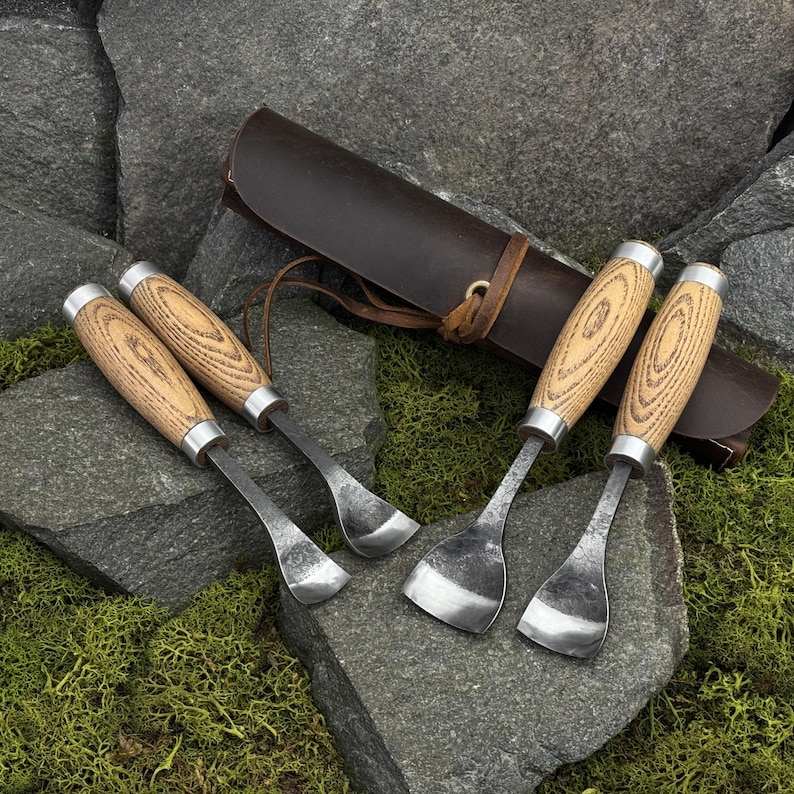 May include: A set of four woodworking tools with light-colored wood handles and silver metal ferrules. Two tools feature curved blades, while two have wider, flat-edged blades. A dark brown leather roll-up case with a leather tie is included.