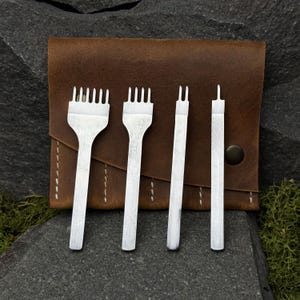 May include: A set of four silver leatherworking tools, including pronged punches and a single-prong tool, displayed against a brown leather case. The tools are designed for creating precise holes in leather. The case has a snap closure.