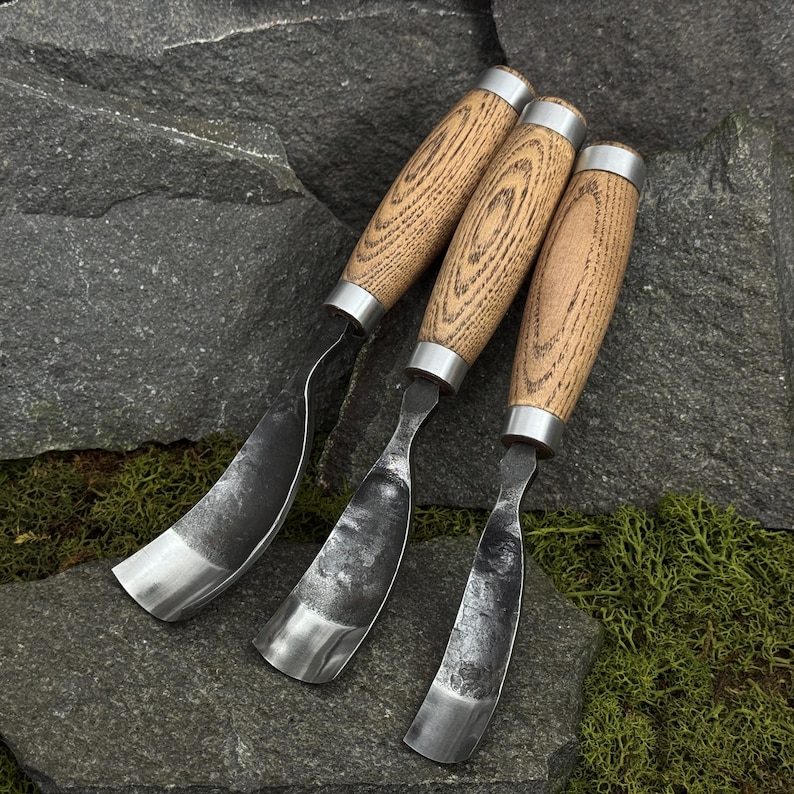 May include: Three hook knives with curved, dark metal blades and light brown wooden handles featuring visible grain patterns. Each tool has a silver-colored metal ferrule connecting the handle to the forged steel blade, designed for woodworking.