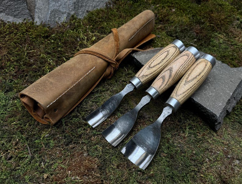 May include: Set of three wood carving tools with wooden handles and a leather roll-up case. The tools have a silver finish and are arranged on a green mossy surface.