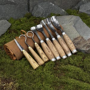 May include: A set of wood carving tools with wooden handles and metal blades. The tools include a knife, hook knives, and various gouges. The tools are arranged next to a brown leather tool roll, set against a backdrop of green moss and stone.