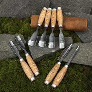 May include: A collection of wood carving chisels with wooden handles and metal blades. The chisels are arranged on a bed of green moss and gray stones, with a brown leather roll in the background. The tools are designed for woodworking and carving.