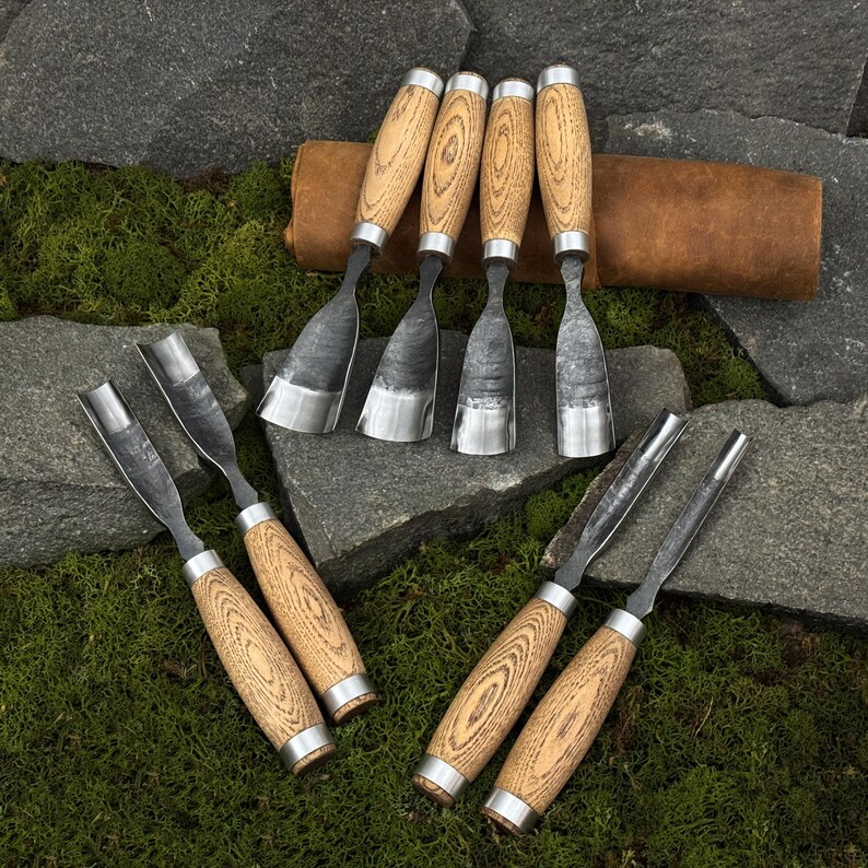 May include: A collection of wood carving chisels with wooden handles and metal blades. The chisels are arranged on a bed of green moss and gray stones, with a brown leather roll in the background. The tools are designed for woodworking and carving.