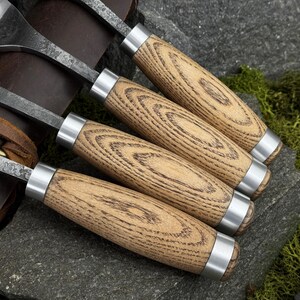 May include: A set of four wood carving tools featuring textured light brown wooden handles with prominent grain patterns, silver metal ferrules, and dark forged steel blades, arranged in a row with a brown leather roll case visible underneath.