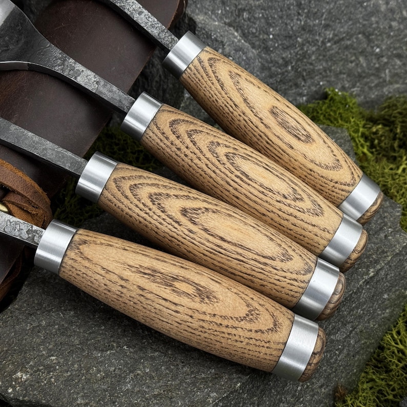 May include: A set of four wood carving tools featuring textured light brown wooden handles with prominent grain patterns, silver metal ferrules, and dark forged steel blades, arranged in a row with a brown leather roll case visible underneath.