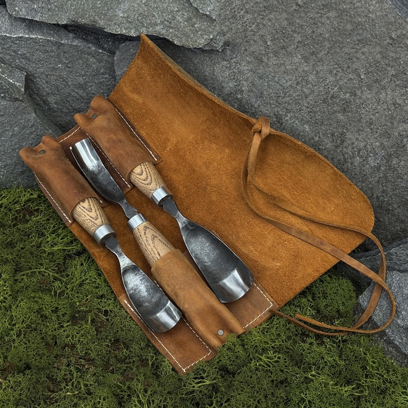 May include: A set of three wood carving gouges with polished metal blades and light-colored wooden handles, secured within a tan leather tool roll featuring stitched pockets and a long leather tie for closure and storage of the carving tools.