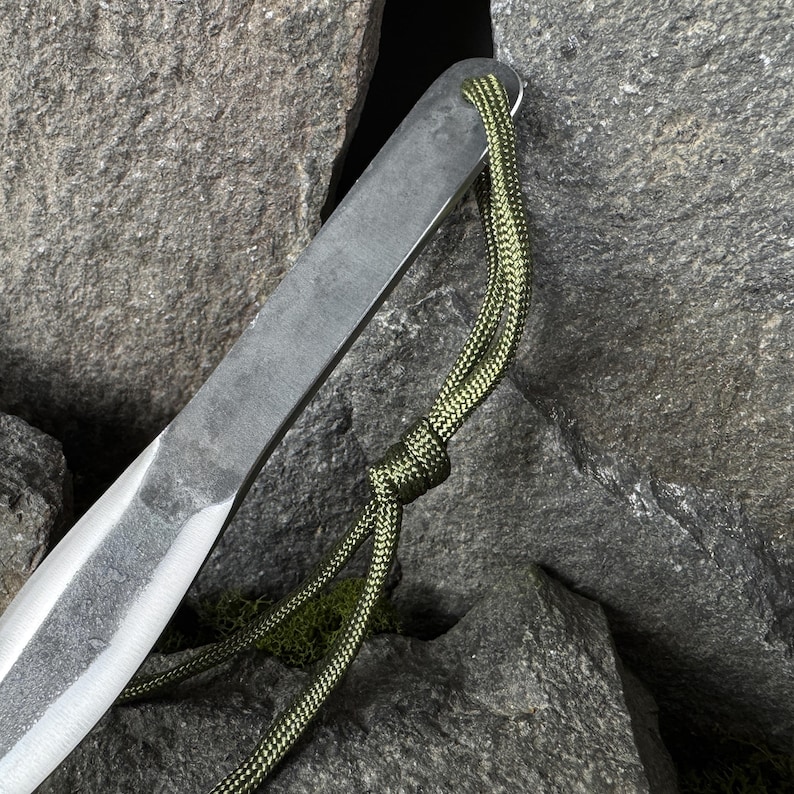 May include: Close-up of a metal tool with a sharpened edge and a green cord attached. The tool has a dark, textured finish and a loop for the cord. The cord is a woven green and black pattern. The background is a collection of gray rocks.