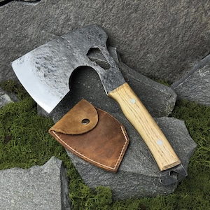 Peut inclure: Une hache en acier avec un manche en bois et un étui en cuir marron. La hache possède un ouvre-bouteille et un trou hexagonal. La lame est argentée et le manche est brun clair.
