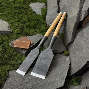 May include: Two hand-forged steel chisels with long, light-colored wooden handles and wide, flat metal blades with sharpened edges, accompanied by a small, rectangular brown leather case with visible stitching along the edges.