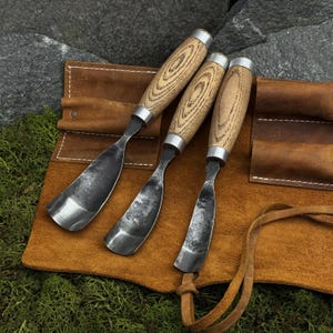 May include: Three wood carving gouges with curved, dark metal blades and light-colored wooden handles featuring visible grain patterns. The tools are arranged on a brown leather roll-up tool pouch with stitched pockets and a leather tie closure.
