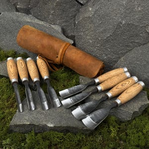 May include: A set of wood carving chisels with wooden handles and metal blades, arranged on a stone surface. A brown leather tool roll is also present. The chisels are designed for woodworking and carving projects.