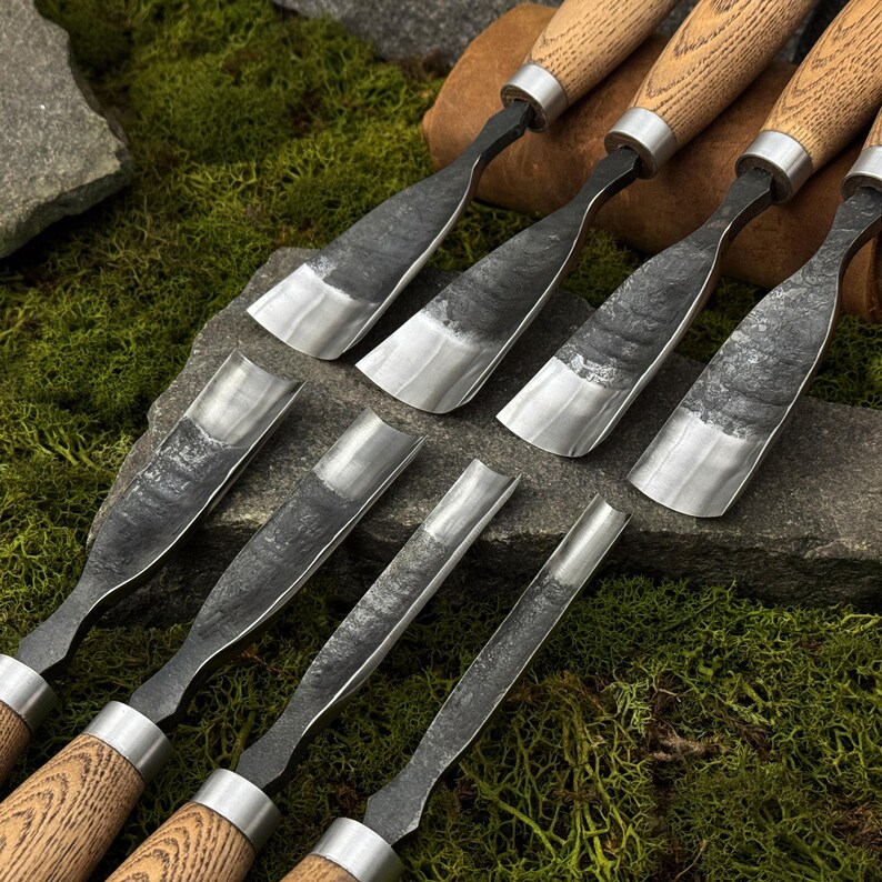 May include: A collection of wood carving chisels with wooden handles and metal blades. The blades have a dark, textured appearance, and the handles are a light brown color. The chisels are arranged on a stone surface with green moss.