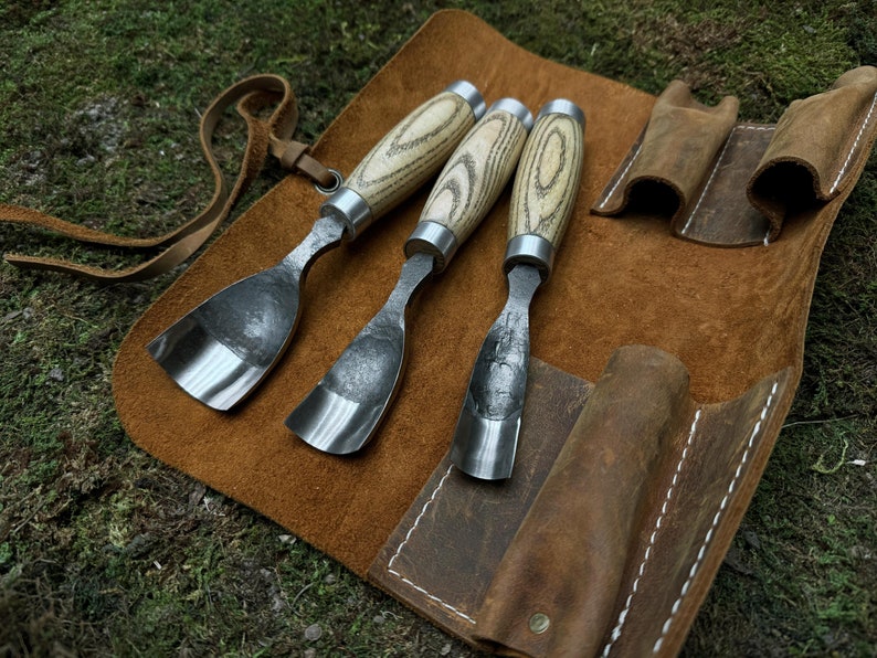 May include: Three wood carving tools with wooden handles and metal blades are rolled up in a brown leather case with white stitching.