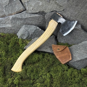 Peut inclure: Hache avec un manche en bois clair et une tête en métal foncé. La hache est accompagnée d'un étui en cuir marron. La hache est posée sur un lit de mousse verte et de roches grises.