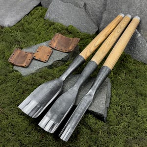 May include: Three wood-handled chisels with metal blades, arranged diagonally on a bed of green moss and gray stones. The chisels have light-colored wooden handles and dark metal blades. Three small, brown leather sheaths are also visible.