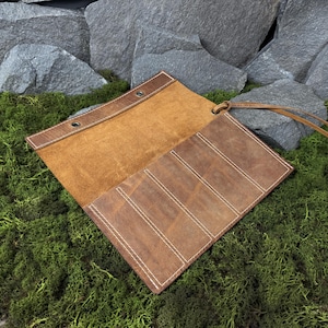 Puede incluir: Estuche enrollable de cuero marrón abierto, con costuras blancas. El estuche tiene una solapa con dos ojales y una correa de cuero. El interior es de un tono marrón más claro. El estuche está sobre un lecho de musgo verde.