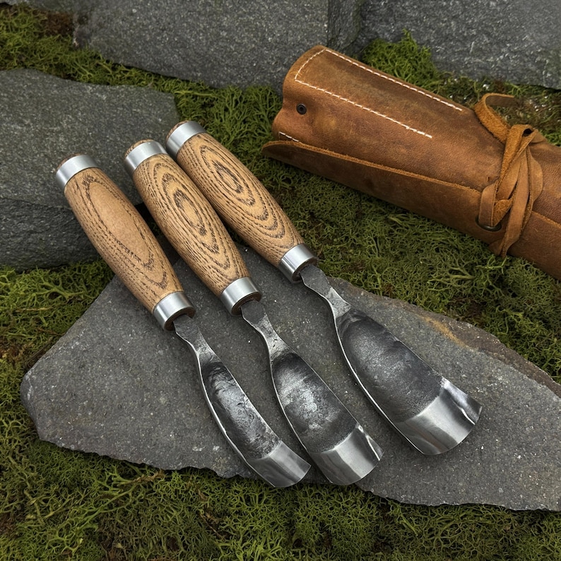 May include: Three wood carving gouges with curved, dark metal blades and light brown wooden handles with visible grain patterns. The tools feature silver-colored metal ferrules connecting the blade to the handle, accompanied by a brown leather roll.