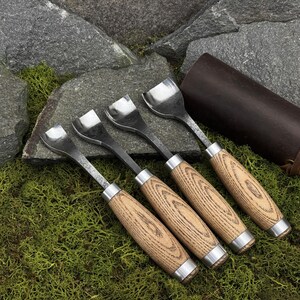 May include: A set of four wood carving gouges with curved metal blades and light brown wooden handles. Each tool features a silver metal ferrule connecting the blade to the handle. The gouges are arranged in a row, showcasing their varying blade widths.
