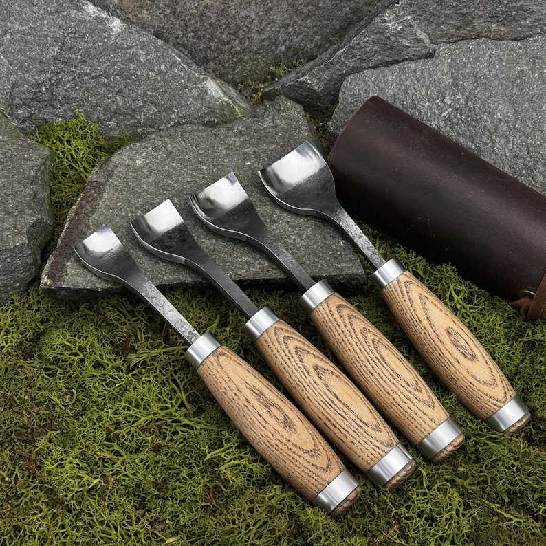 May include: A set of four wood carving gouges with curved metal blades and light brown wooden handles. Each tool features a silver metal ferrule connecting the blade to the handle. The gouges are arranged in a row, showcasing their varying blade widths.