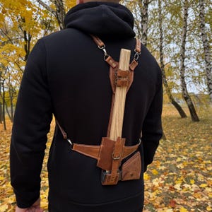May include: A brown leather axe carrier with a wooden axe. The carrier has adjustable straps and a small pouch. The axe head is secured in a leather sheath. The carrier is designed to be worn on the back.