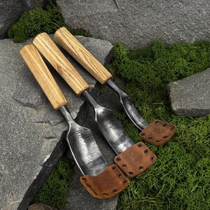 May include: Three hand tools with light brown wooden handles and dark forged metal blades. Each blade features a curved shape and is protected at the tip by a brown leather cover secured with small metal rivets, creating a functional set.