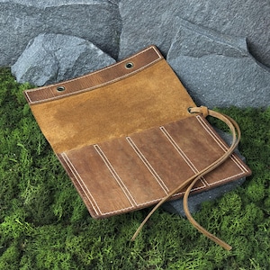 May include: Open leather roll-up case in shades of brown. The case is made of distressed leather with white stitching and a leather tie closure. The interior is a lighter tan colour. The case is designed to hold multiple items.