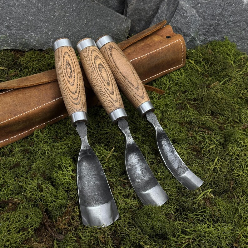 May include: Three wood carving gouges with curved metal blades and light brown wooden handles featuring visible grain patterns. Each tool has a silver metal ferrule connecting the handle to the steel blade. The tools are arranged side by side.