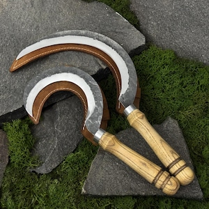 May include: Two curved sickles with forged metal blades and light-coloured wooden handles. Each blade is partially encased in a brown leather sheath with visible stitching. The tools feature a textured, dark metal finish on the curved edges.