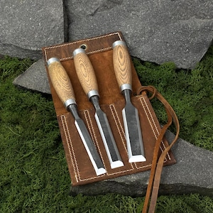 May include: A set of three wood chisels with light brown wooden handles and silver metal ferrules, stored in a brown leather tool roll with white stitching and a leather tie closure. The chisels feature flat, sharp metal blades.