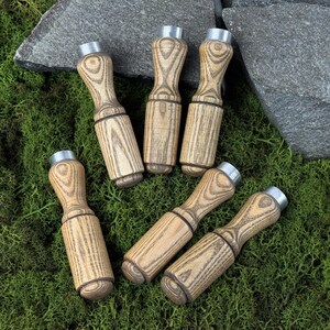 Peut inclure: Six appeaux à canard en bois clair, avec un motif de grain. Chaque appeau a un dessus en métal argenté et une bande marron foncé. Ils sont disposés sur de la mousse verte. Idéal pour la chasse.