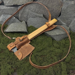 May include: A brown leather axe with a wooden handle, secured in a matching leather sheath. The sheath has a strap for carrying. The axe is resting on a bed of green moss, with a backdrop of gray rocks.