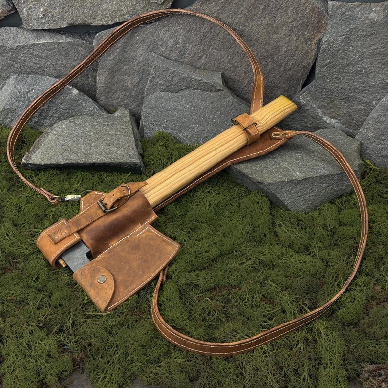 May include: A brown leather axe with a wooden handle, secured in a matching leather sheath. The sheath has a strap for carrying. The axe is resting on a bed of green moss, with a backdrop of gray rocks.