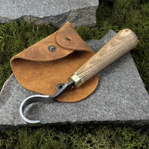 May include: A wood carving hook knife with a curved blade and a wooden handle, resting on a gray stone surface. The knife is accompanied by a brown leather pouch with a snap closure. The handle has a brass ferrule.