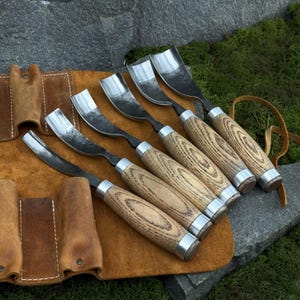 May include: A set of five wood carving gouges with curved steel blades and light brown wooden handles, each featuring a metal ferrule. The tools are arranged in a row on a tan leather roll-up tool pouch with stitched pockets and a leather tie.