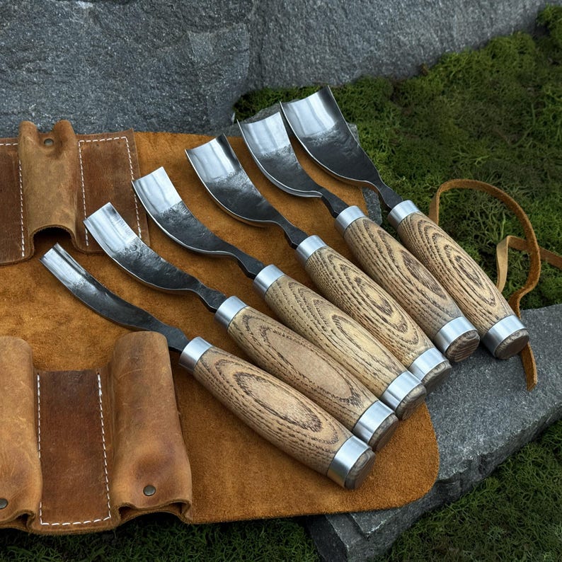 May include: A set of five wood carving gouges with curved steel blades and light brown wooden handles, each featuring a metal ferrule. The tools are arranged in a row on a tan leather roll-up tool pouch with stitched pockets and a leather tie.