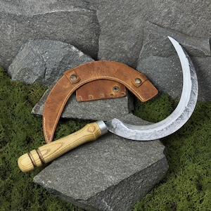 May include: A hand-forged sickle with a curved, textured blade and a wooden handle. The sickle is accompanied by a brown leather sheath. The tools rest on a gray stone surface with green moss.