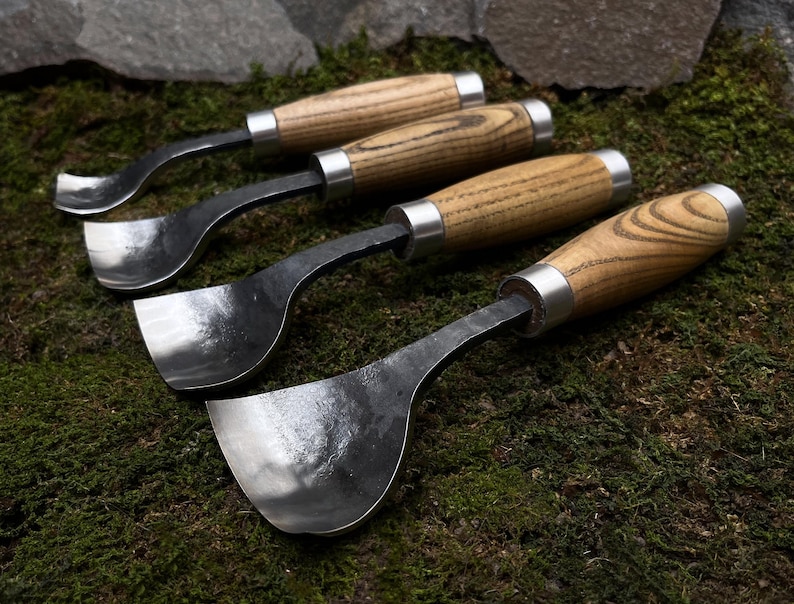 May include: Four wood carving tools with curved blades and wooden handles. The tools are arranged in a row on a bed of green moss.