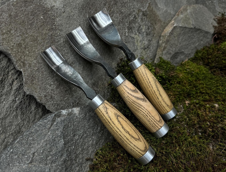 May include: Three wood carving tools with wooden handles. The tools have a curved blade and a metal ferrule. The handles are made of light brown wood with dark brown streaks.