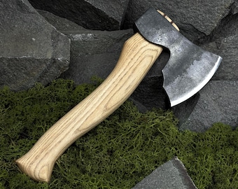 May include: A handcrafted axe with a light-colored wooden handle and a dark, forged steel head. The axe head has a sharp, curved blade. The handle is made of wood and has a smooth, ergonomic design. The axe is designed for chopping and splitting wood.