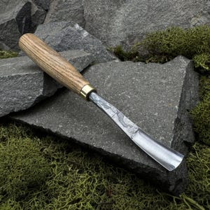 May include: A woodworking chisel with a wooden handle and a metal blade. The handle is light brown with a brass ferrule. The blade is silver and has a curved cutting edge. The chisel is resting on a gray stone.