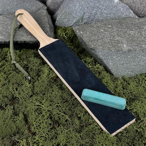 Puede incluir: Un asentador de madera con una superficie de cuero negro y una piedra de afilar verde claro. El asentador tiene un mango de madera clara y un cordón verde para colgar. La piedra de afilar es rectangular.