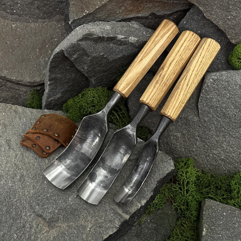 May include: Three metal gouges with curved, dark grey steel blades and light brown wooden handles, arranged side by side. A small, folded brown leather piece with metal rivets is positioned to the left of the metal tools.