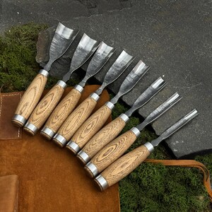 May include: A set of eight wood carving chisels with wooden handles and metal blades. The handles have a light wood grain pattern and are accented with silver-colored metal rings. The chisels are arranged on a brown leather surface.