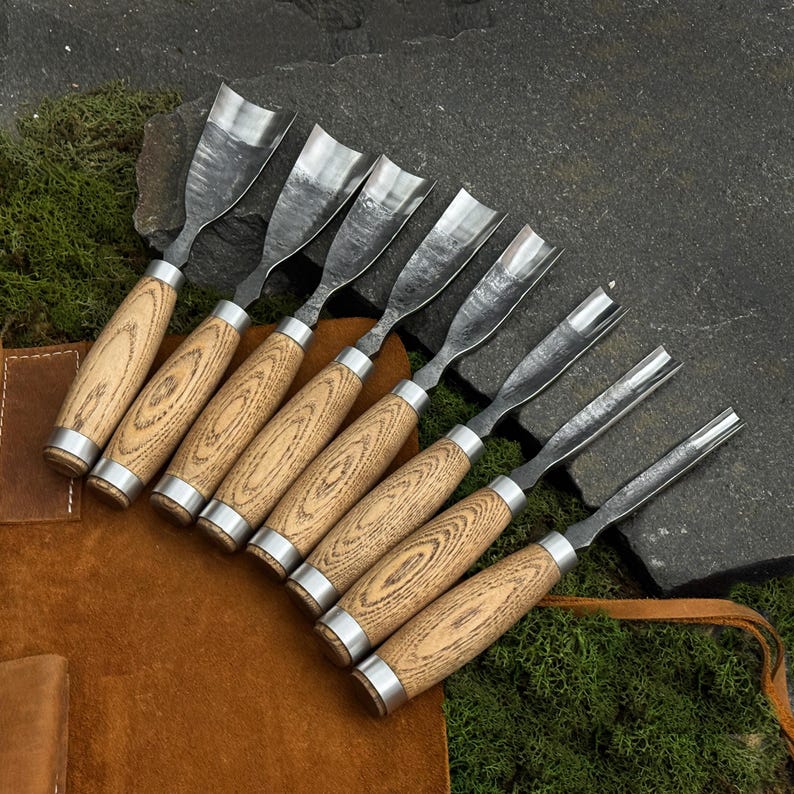 May include: A set of eight wood carving chisels with wooden handles and metal blades. The handles have a light wood grain pattern and are accented with silver-colored metal rings. The chisels are arranged on a brown leather surface.