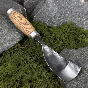 May include: A hand-forged metal spoon gouge featuring a curved, hammered steel blade and a cylindrical wooden handle with a visible grain pattern, secured by two silver-toned metal ferrules at the top and bottom of the grip.