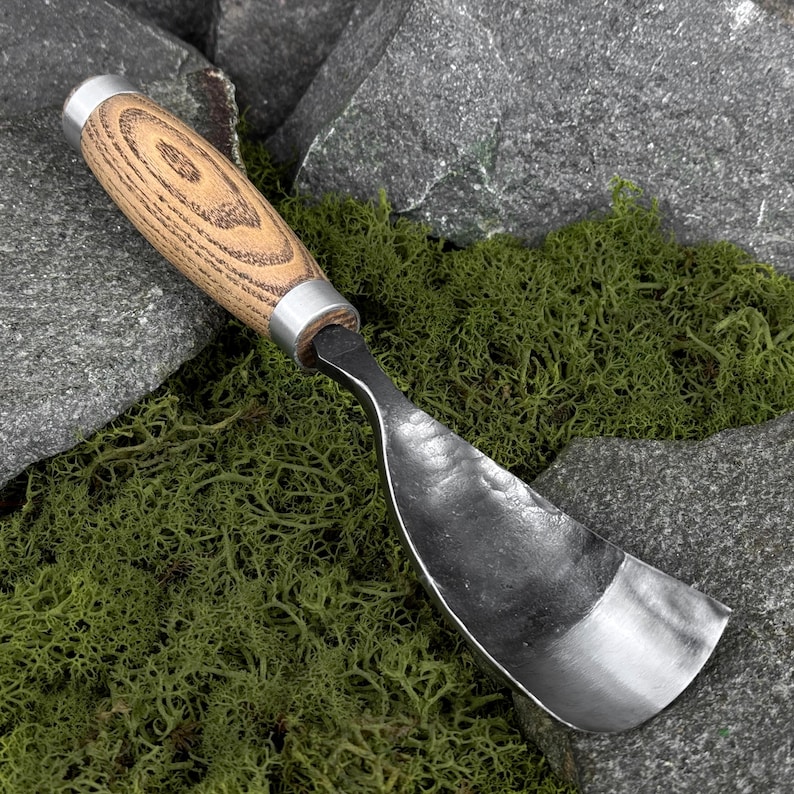 May include: A hand-forged metal spoon gouge featuring a curved, hammered steel blade and a cylindrical wooden handle with a visible grain pattern, secured by two silver-toned metal ferrules at the top and bottom of the grip.
