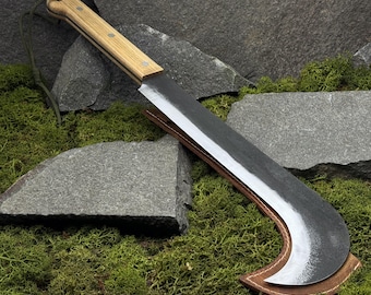 May include: A large, curved machete with a light wood handle and a dark, textured blade. The blade is partially sheathed in a brown leather cover. A green cord is attached to the handle. The machete rests on a bed of green moss and gray stones.