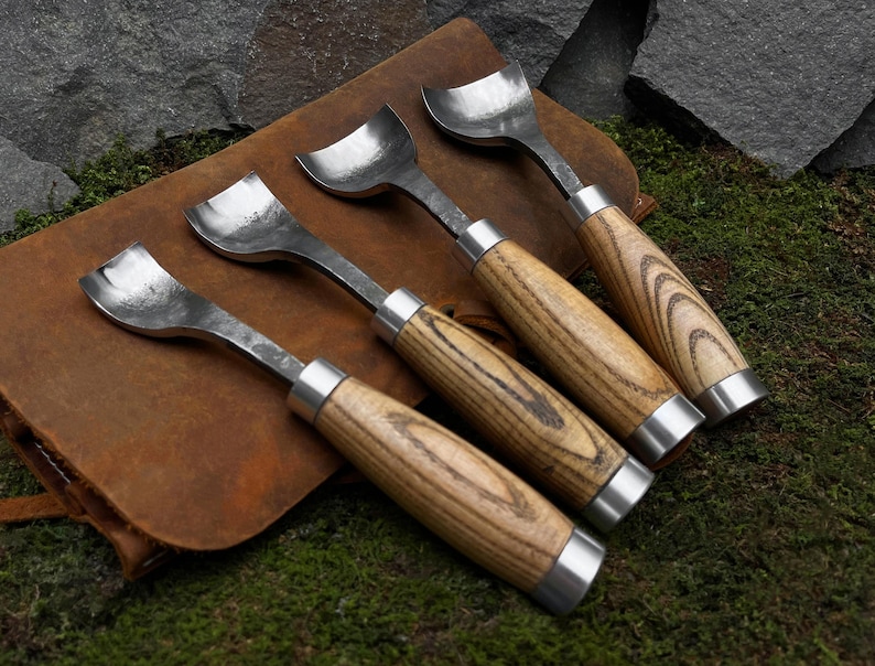 May include: Four wood-handled carving tools with metal blades. The tools have different shaped blades and are arranged on a brown leather surface.