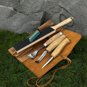 May include: A set of three wood carving tools with light-coloured wooden handles, a rectangular wooden sharpening strop with a black abrasive surface, and a small green polishing compound block, all arranged on a tan leather tool roll.